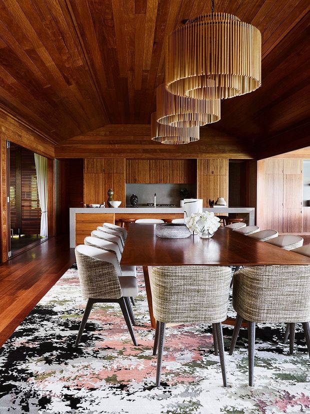 A warm, richly detailed open-plan dining and kitchen interior. A large dark timber dining table is surrounded by upholstered dining chairs and positioned over a patterned area rug. The room features extensive dark timber wall and ceiling panelling, timber flooring, integrated timber cabinetry, a pale stone kitchen island, and three cascading cylindrical slatted pendant lights. Full-height glazing and timber-framed openings are visible at the sides.
