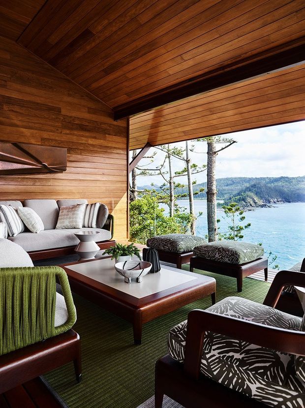 A richly timber-lined coastal living room opening onto a broad ocean and bay view. The room features a pitched timber ceiling, horizontal timber wall panelling, a large open glazed frontage, a grey sofa, dark wood-framed lounge chairs with patterned upholstery, a timber coffee table, and a deep green textured rug. The setting feels warm, refined, and strongly connected to the surrounding coastal landscape.