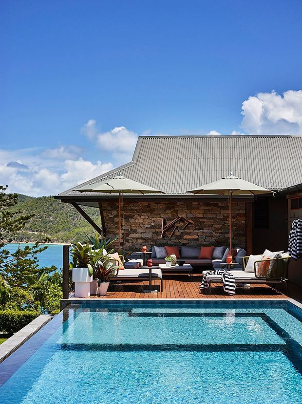 A bright outdoor terrace with an infinity-edge swimming pool overlooking a vivid blue body of water and mountainous terrain. The terrace features timber decking, a dark stone feature wall, outdoor lounge seating, umbrellas, and tropical planting. A large grey corrugated metal roof forms the structure behind the entertaining area.