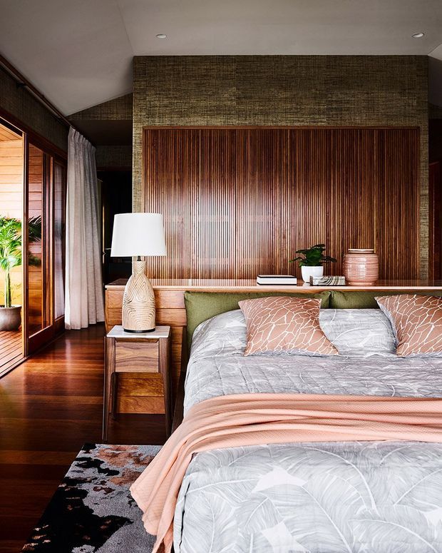 Warm, richly detailed bedroom interior with a low upholstered bed, layered grey and blush bedding, patterned cushions, and integrated wooden bedside storage. The room features a dramatic vertical timber slat feature wall framed by darker textured wall panels, polished dark timber flooring, floor-to-ceiling timber-framed glazing with curtains, and layered ambient lighting. Tropical greenery is visible at the glazing, reinforcing the natural, resort-like atmosphere.