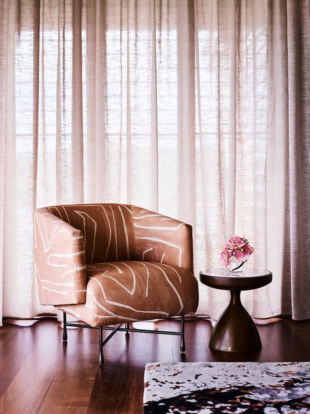 A styled interior vignette featuring a sculptural upholstered lounge chair in a warm terracotta and brown abstract pattern, positioned beside a dark wood pedestal side table with a small floral arrangement. Sheer floor-to-ceiling curtains filter soft daylight, with polished dark timber flooring and a patterned area rug completing the setting. The scene has a refined, warm, softly dramatic feel.