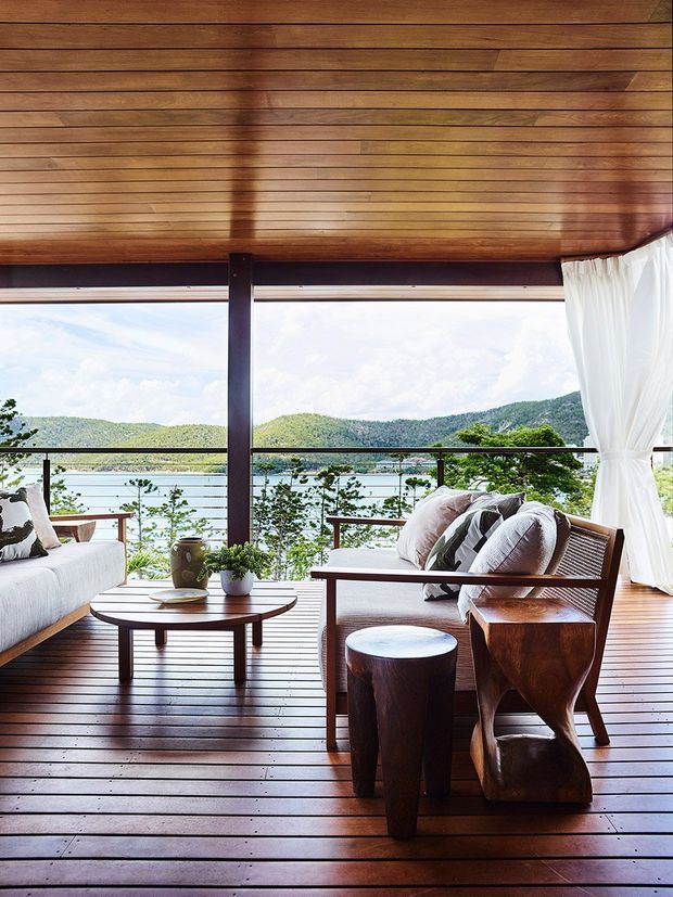 An elevated, open-air living lounge with a dark timber slatted ceiling and matching timber deck floor. The space features a white upholstered sofa, wooden-framed lounge seating with neutral cushions, a low coffee table, sculptural timber stools and potted plants. Full-height openings with sheer white curtains overlook a tranquil lake and wooded hills, creating a relaxed indoor-outdoor setting.