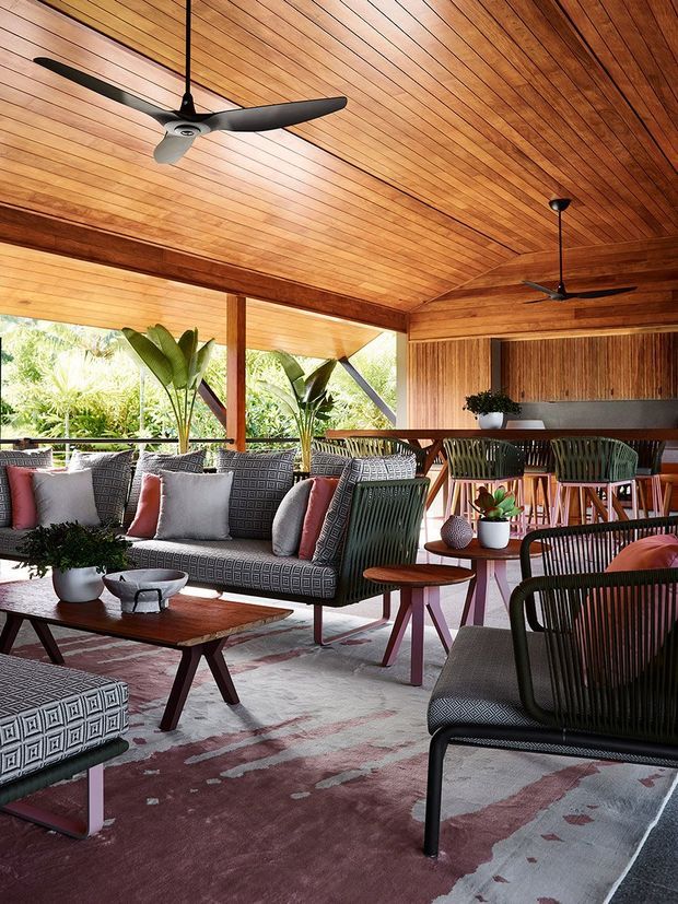 A stylish semi-enclosed outdoor lounge or entertaining area with extensive warm timber lining across the ceiling and walls, exposed timber beams and posts, and open glazing to lush vegetation. The space is furnished with woven lounge seating, patterned cushions, timber coffee tables, bar-height dining seating, potted plants, ceiling fans, and a large muted patterned rug. The overall atmosphere is warm, relaxed, tropical, and refined.