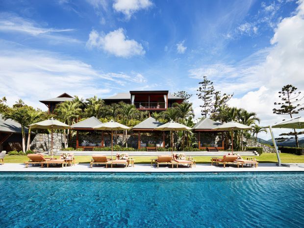 A bright tropical resort-style exterior with a large turquoise swimming pool in the foreground, rows of loungers and pale parasols along the pool deck, lush palm landscaping, and a dark timber-clad multi-level building behind.