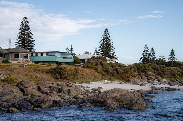 A low-rise coastal residential property viewed from the rocky shoreline, with a long modernist building, broad horizontal glazing, pale green and grey exterior surfaces, and a gently sloping roof. The house sits amid coastal scrub and tall Norfolk Island pines, overlooking a rocky beach and blue water under a partly cloudy sky.