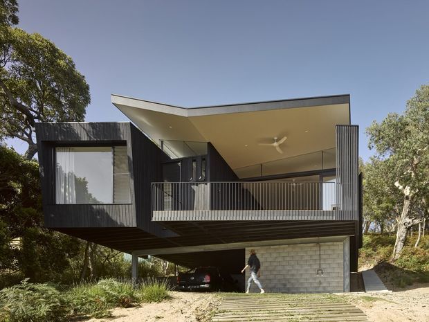 Exterior view of a contemporary elevated home set among mature trees on a sloping site. The house features dark vertical cladding, a projecting upper-level volume, broad roof overhangs, open balcony railings, extensive glazing, and a ground-level concrete-block garage or undercroft.