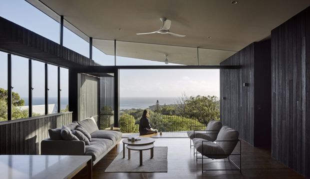 A contemporary living room with floor-to-ceiling glazing opening toward a coastal landscape and ocean view. The room features dark vertical timber wall cladding, a pale ceiling with ceiling fans, a large grey sofa, sculptural lounge chairs, nesting coffee tables, and a neutral rug. The open, glazed composition creates a strong indoor–outdoor connection and frames the elevated coastal outlook.