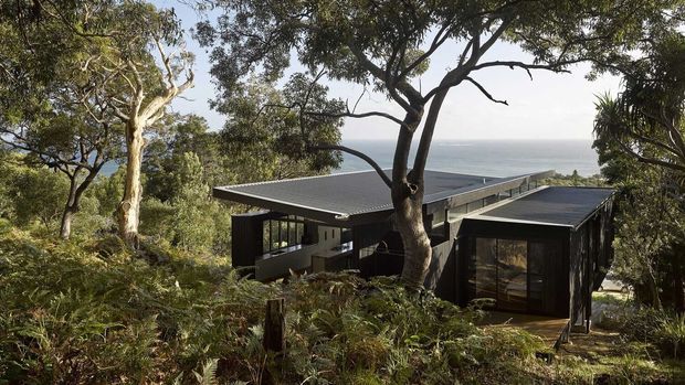 A modern, low-profile house nestled among dense coastal bushland, with dark cladding, extensive glazing, and broad flat or very low-pitched roof planes. The home appears elevated on a sloping site, with views toward the ocean visible beyond the trees.