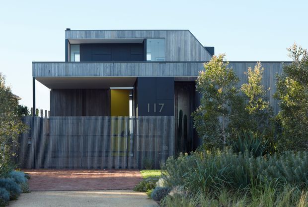 Front exterior view of a contemporary multi-level residence with strong horizontal and vertical lines, dark recessed openings, and weathered grey timber cladding. A screened entry courtyard, brick-paved driveway, and dense native-style landscaping frame the facade. The upper level includes a projecting balcony or terrace, while a bright yellow entry door provides a small accent.