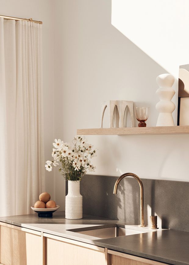 Warm, minimalist kitchen vignette with pale timber cabinetry, a dark stone benchtop and matching backsplash, an undermount sink with a curved brass tap, and open shelving displaying neutral-toned decor. Soft sunlight casts strong geometric shadows across the light plaster walls, with linen curtains and natural floral styling adding an organic touch.