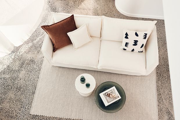 Overhead view of a bright, minimalist living room vignette with a curved cream sofa, neutral textured rug, small round side table, dark glass coffee table, and a restrained mix of cream, brown, and graphic black-and-white cushions. The space has a soft, contemporary and calm aesthetic.