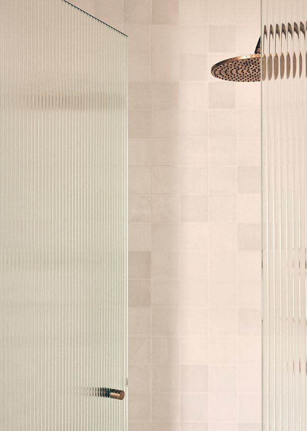 Close-up view of a contemporary shower enclosure. Ribbed or fluted translucent glass panels frame a pale square-tiled shower wall, with a large round brushed metal rainfall showerhead and a small matching control fitting visible. The palette is soft white and warm neutral with subtle metallic accents.