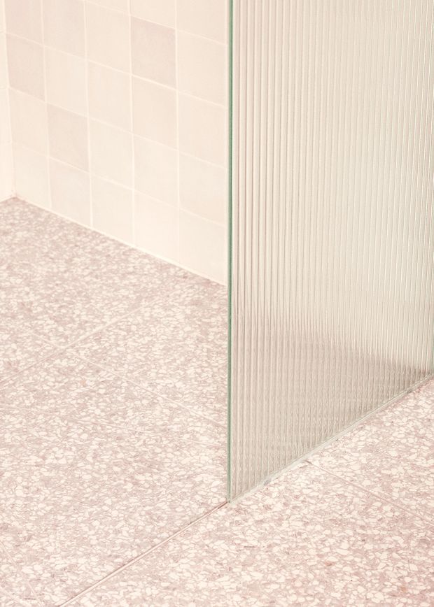 Close-up architectural interior detail showing pale pink terrazzo-style flooring, pale blush square wall tiles, and a vertically ribbed translucent glass partition with a slim frame. The image is minimalist and softly tonal, focused on material junctions and surface textures.