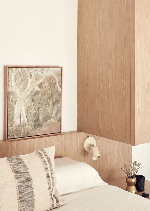 Warm, softly styled bedroom corner featuring a bed with neutral linen bedding and a striped woven cushion. Vertical light timber wall panelling and matching built-in joinery frame the bed, with a small round wall-mounted reading light. A framed muted botanical artwork, bedside vase with dried branches, and dark accessories add restrained decorative detail.