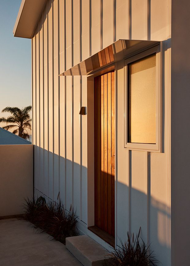 Close exterior view of a contemporary residential entry, featuring vertically ribbed pale cladding, a warm timber pivot door, a narrow frosted-glass sidelight or window, and a projecting timber-and-metal canopy. A concrete path and simple planted border lead to the entrance, with strong low-angle sunlight creating pronounced shadows.