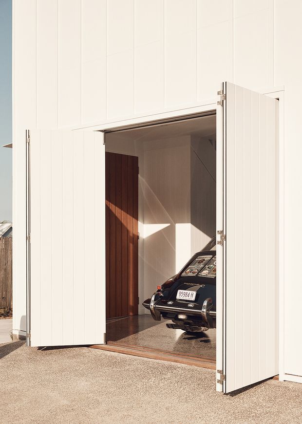 Exterior view of a minimalist white-clad garage with large double doors open, revealing a vintage dark-colored car parked inside. The garage has vertical panel cladding, a smooth concrete floor, warm timber wall lining at the rear, and strong sunlight creating angular shadows.