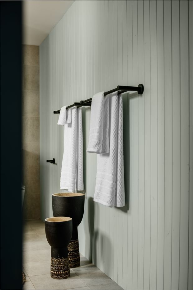A narrow, moody bathroom with white vertical wall panelling, pale stone or concrete-look floor and wall tiles, black metal towel rails and fixtures, and two decorative black vessel basins with patterned lower sections. White textured towels hang prominently on the rails, with a mirror reflecting the space.