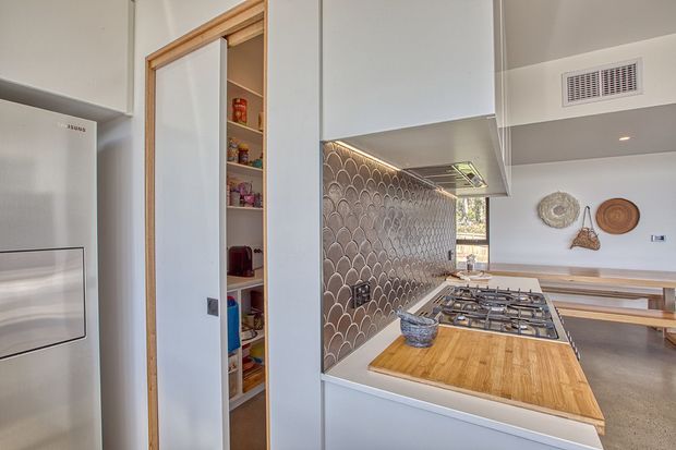 A contemporary galley-style kitchen viewed toward a dining area, with white built-in cabinetry, a tall integrated refrigerator, a concealed walk-in pantry, timber-framed pantry door, timber benchtop section, patterned scallop-shaped tile splashback, and a stainless steel gas cooktop. Polished concrete flooring and decorative woven wall pieces are visible in the adjoining dining space.