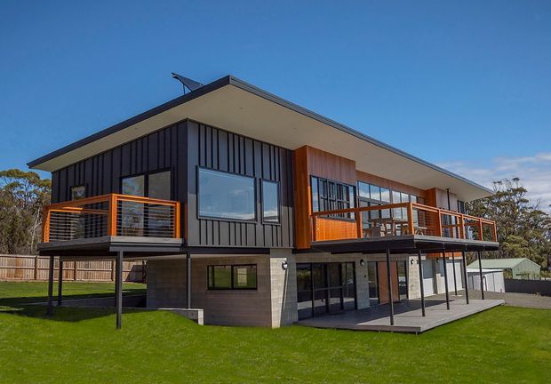 Exterior photograph of a contemporary two-storey home on a gently sloping landscaped site. The building has a broad low-pitched roof overhang, dark vertical cladding, warm timber accents, extensive glazing, elevated upper-level balconies with orange-brown metal balustrades, and a covered lower-level terrace.