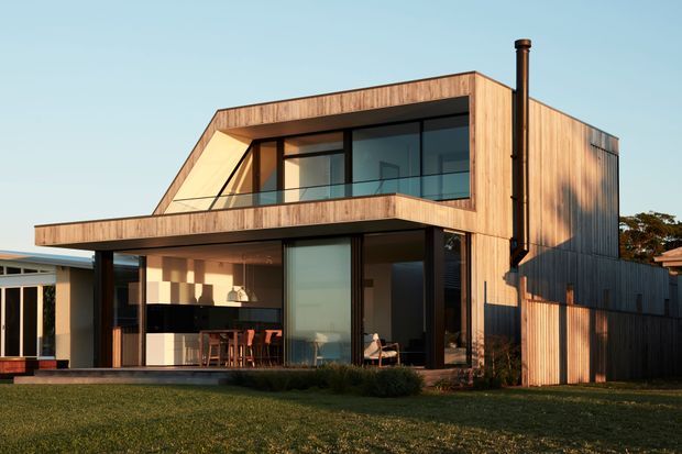 Exterior view of a contemporary two-storey residence with a strong geometric form, extensive dark-framed glazing, and warm vertical timber cladding. The upper level projects over a covered ground-floor terrace and open-plan living area, while a tall black flue rises beside the house. The home sits in a suburban landscaped setting with lawn and neighbouring buildings visible.