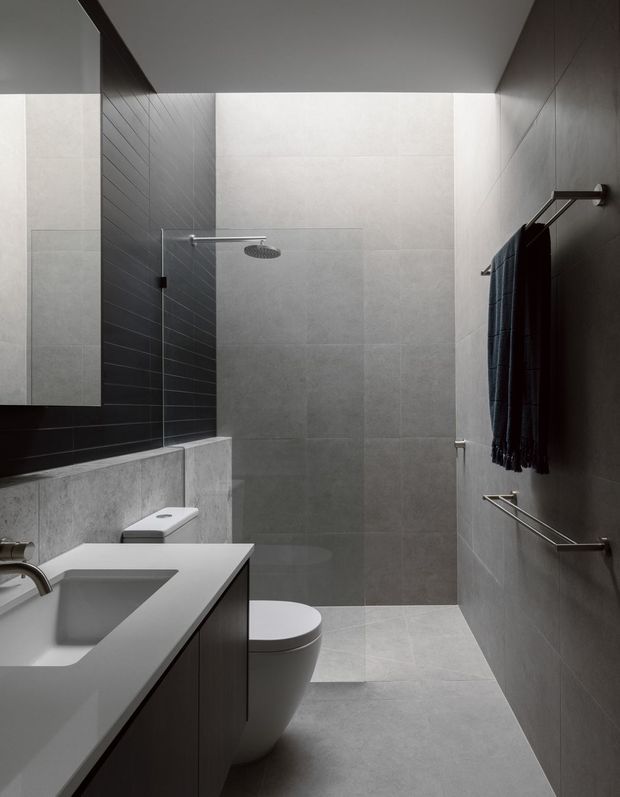 A compact contemporary bathroom with a walk-in shower, wall-mounted rainfall showerhead, toilet, and floating vanity with integrated rectangular basin. Large-format grey porcelain or ceramic tiles cover the floor and walls, with a darker charcoal tiled shower feature wall. Minimal chrome towel rails and dark textiles create a restrained, monochromatic interior.