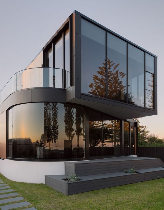 Exterior view of a contemporary multi-level residence with a dramatic cantilevered upper volume, extensive dark-framed floor-to-ceiling glazing, a curved balcony, and dark horizontal cladding. Broad timber-look steps and integrated planter boxes lead through a landscaped lawn to the glazed ground floor. Reflections show surrounding trees and the warm evening sky.