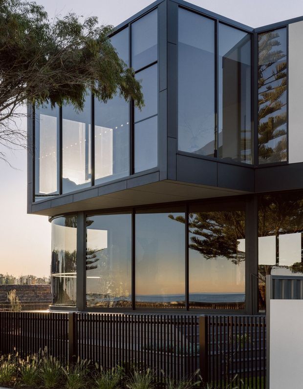Exterior view of a contemporary, multi-level residence with projecting upper volumes, dark framed floor-to-ceiling glazing, and a restrained dark facade. The building overlooks a visible body of water, with vertical metal fencing and coastal vegetation in the foreground. Strong reflections of trees and the surrounding landscape appear in the glazing.