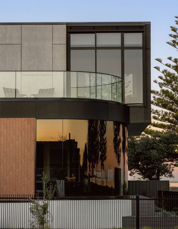 A close exterior view of a contemporary multi-storey residence with a strong geometric composition. The facade combines large pale rectangular cladding panels, dark metal-framed glazing, a curved glass balustrade, dark curved cladding, warm vertical timber boards, and reflective curved ground-floor windows. A balcony and landscaped residential setting are visible, with a restrained palette of charcoal, grey, timber brown, and clear glass.
