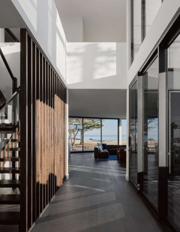 A bright, double-height interior hallway or landing leading into an open living room, with a dark tiled floor, white walls, extensive floor-to-ceiling glazing, and a dramatic timber vertical-slat screen enclosing the stairway. A dark sofa is visible in the living area beyond, with a coastal landscape seen through the windows.