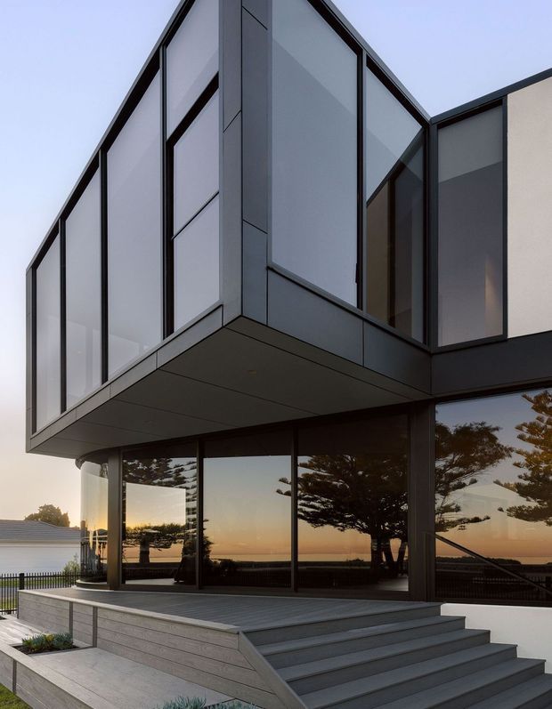 Exterior view of a sharply angular contemporary house with a dramatic glazed upper-level projection and dark metal-framed floor-to-ceiling windows. The home features pale grey cladding, a projecting box-like form, a broad outdoor deck with wide steps, and strong reflections of trees and a coastal-looking horizon in the glazing. The composition emphasizes the building facade and cantilevered geometry at dusk.