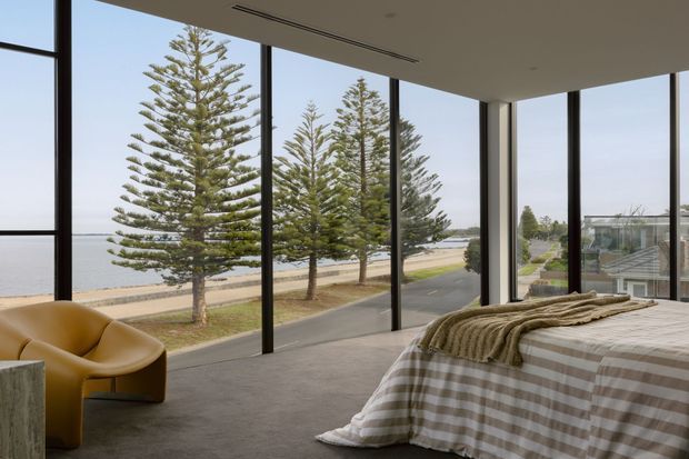 A contemporary bedroom with floor-to-ceiling corner glazing overlooking a waterfront promenade lined with Norfolk Island pines. The room contains a low-profile bed dressed in a neutral checked coverlet and layered throws, a sculptural mustard lounge chair, soft carpet flooring, and a minimalist recessed ceiling. Black-framed glazing creates an airy connection to the coastal landscape.