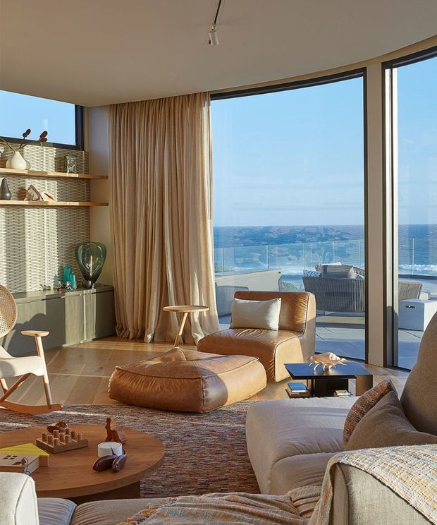 A warm, mid-century-inspired living room with expansive floor-to-ceiling glazing overlooking the ocean. The room features a curved corner window, full-height beige curtains, timber shelving and cabinetry, a textured patterned wall, leather lounge chair and ottoman, a large neutral sofa, timber coffee tables, and a woven area rug. An outdoor terrace with glass balustrading and seating is visible beyond the glazing.