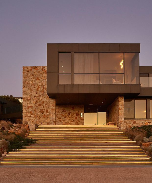 Twilight exterior view of a contemporary multi-level residence. Broad illuminated steps lead up to a recessed entry beneath a projecting upper volume. The facade combines warm, irregular natural stone walls with dark metal or panel-clad sections and large glazed windows. Low ornamental planting borders the stairway, while warm linear step lighting creates a dramatic, welcoming approach.