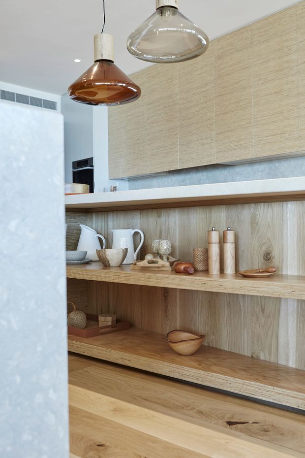 Close-up view of a refined kitchen with extensive light timber open shelving and matching timber flooring. Pale wood cabinetry occupies the upper wall, while a white benchtop and grey stone-look backsplash provide contrast. Two sculptural glass pendant lights hang above, and the shelves display neutral-toned ceramics, wooden utensils, and pantry-style objects. The composition has a warm, natural, curated contemporary feel.