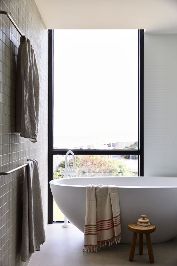 A bright contemporary bathroom featuring a freestanding white oval bathtub positioned beside a large floor-to-ceiling window. The left wall is clad in small grey rectangular tiles with slim metal towel rails and hanging neutral towels. A minimalist curved metal bath tap, pale floor, dark window frames, and a small wooden stool complete the calm, refined space.