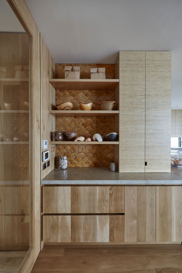 Close-up view of a warm, wood-lined kitchen or bar joinery area. Floor-to-ceiling timber cabinetry frames open shelves lined with a distinctive scalloped terracotta-toned tile backsplash, displaying bowls and ceramics. A stone benchtop sits above timber drawers and cupboards, with a tall integrated cabinet on the right. A glazed timber-framed opening or reflective cabinet is visible on the left, and part of the adjacent kitchen is seen through the opening on the right.