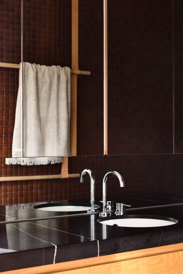 Close-up of a sophisticated bathroom vanity with a dark tiled wall, polished black stone countertop, twin white inset basins, and chrome twin-arched faucets. A warm timber towel rail and light neutral towel provide contrast, while reflective surfaces and strong horizontal lines create a refined, dramatic composition.