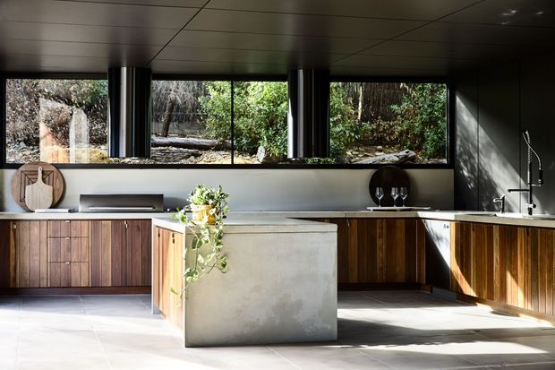 A refined contemporary kitchen with a large concrete island, dark timber cabinetry, pale stone or concrete benchtops, and expansive horizontal windows overlooking a garden and rocky landscape. The space has a dark paneled ceiling, integrated stainless-steel rangehoods, a minimal sink area, and strong natural light creating a warm yet dramatic atmosphere.