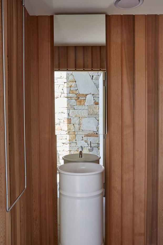 A compact bathroom viewed through a narrow opening, featuring a white cylindrical pedestal basin with wall-mounted faucet, a second basin visible behind it, vertical warm timber wall panelling, a small window with a timber blind, and a rough pale natural-stone wall outside or behind the glazing. The space has a restrained, refined contemporary character with strong natural materials and concealed detailing.