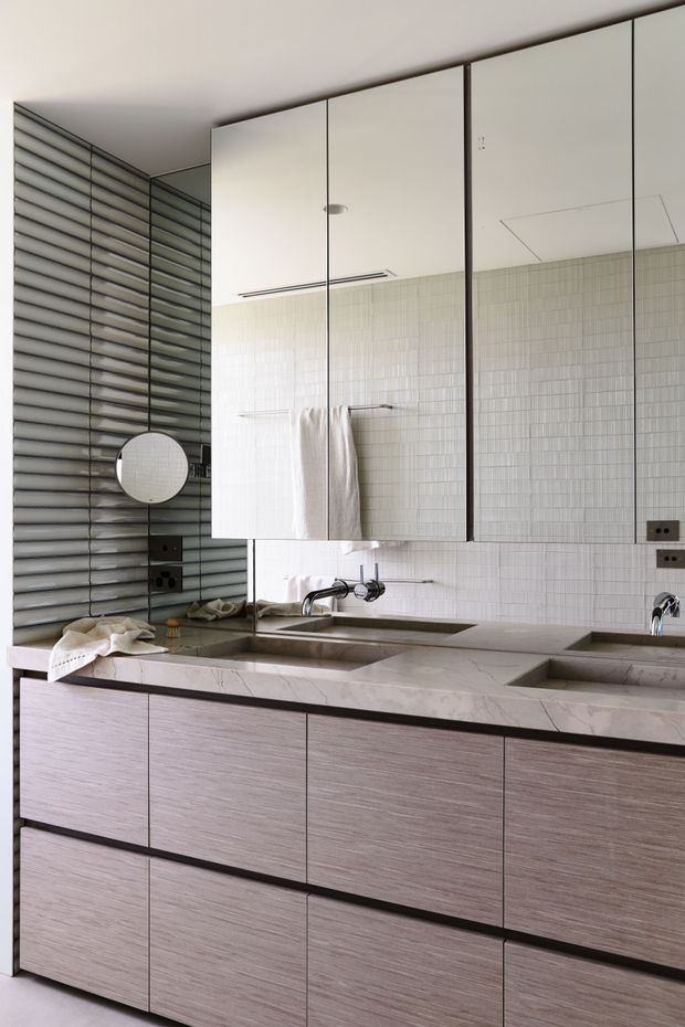 A refined contemporary bathroom vanity with a long double-basin stone countertop, integrated sinks, wall-mounted chrome taps, extensive mirrored upper cabinetry, and timber-look lower drawers. The walls feature small pale rectangular tiles, while the side wall has dark horizontal ribbed tiles or panels. A round shaving mirror, towels, recessed lighting, and dark electrical fittings are visible.