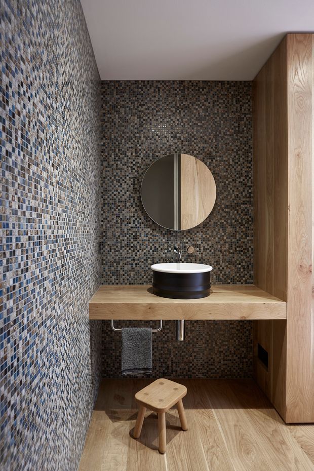 Compact contemporary bathroom with a wall-mounted timber vanity, white vessel basin, black cylindrical base, round mirror, and full-height small-format mosaic tile walls in muted grey, brown, beige, and blue tones. A timber storage cabinet occupies the right side, with timber-look flooring and a small wooden stool beneath the vanity.