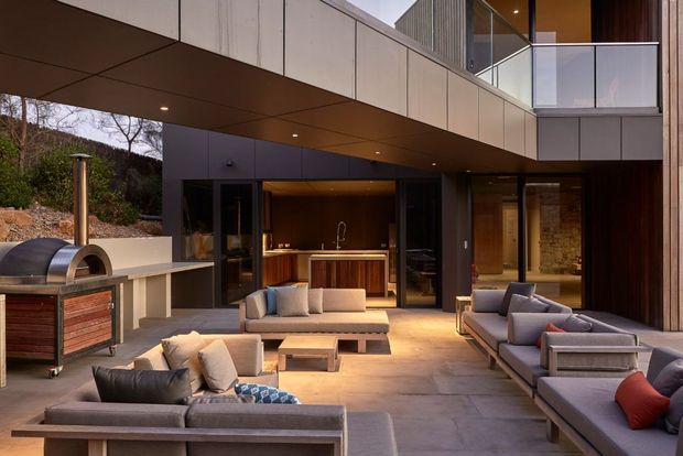 A sophisticated modern outdoor entertaining courtyard beneath a cantilevered upper storey. The space includes a built-in outdoor kitchen/bar with timber cabinetry, a wood-fired pizza oven, extensive contemporary lounge seating, concrete paving, dark framed glazing, and warm timber and stone wall accents. The architecture features strong horizontal lines, a projecting balcony, and a sheltered open-air living arrangement.