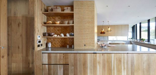 A warm contemporary kitchen with extensive vertical-grain timber cabinetry, a large island with a pale stone benchtop, open timber shelving displaying crockery, and a textured brown backsplash. Pendant lights hang above the island, while full-height glazing brings in natural light and views beyond.