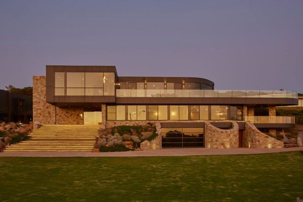 A large contemporary multi-level residence photographed at dusk, featuring extensive warm-lit glazing, broad horizontal balcony terraces, curved upper-level roofline, and extensive natural stone cladding. A broad illuminated staircase leads through landscaped planting to the entrance, while a garage is integrated into the lower stone-clad level.