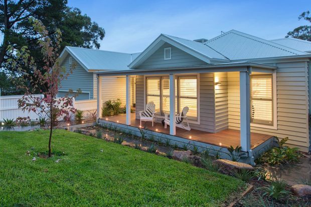 Exterior view of a single-storey weatherboard house at dusk, featuring a covered timber veranda, multiple gabled roof sections, pale grey horizontal cladding, white-framed windows with shutters, and warm interior lighting. The home is set within a landscaped suburban garden.