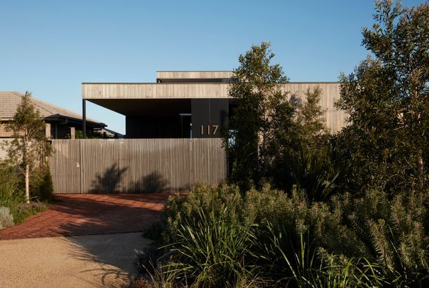 Street-facing view of a contemporary single-storey residence with a boxy flat-roof form, extensive horizontal timber-clad fascia and walls, a dark recessed entry, and a tall timber privacy fence. A red-toned paved driveway and native-style landscaped planting frame the facade.