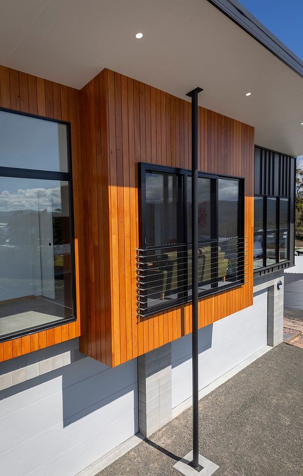 Exterior architectural view of a contemporary elevated house with warm vertical timber cladding, black-framed glazing, a covered soffit, and a black metal balcony or window guard. The lower level uses painted concrete blockwork, with a sloping site and distant landscape visible through the glazing.