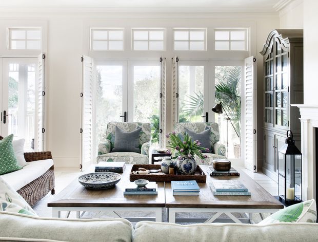 Bright, refined living room with a large upholstered sofa, two patterned armchairs, a substantial wooden coffee table, and extensive white-framed glazed doors and windows with shutters. Built-in or architectural shelving and a white fireplace surround are visible on the right, with layered neutral and soft green furnishings creating an airy coastal feel.