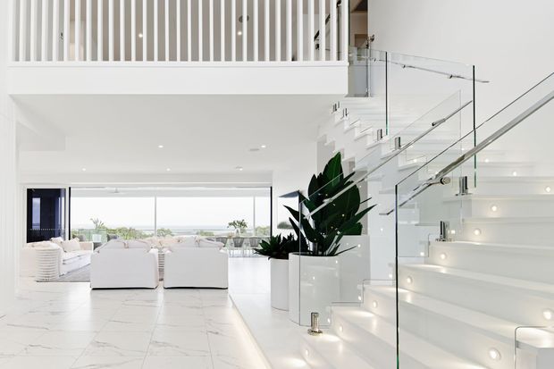 Bright, spacious contemporary open-plan interior featuring a white living area with large floor-to-ceiling glazing, white sofas, marble-look tiled flooring, sculptural indoor plants, and a broad illuminated staircase with frameless glass balustrades. A mezzanine or upper-level landing with white vertical balustrade detailing is visible above.