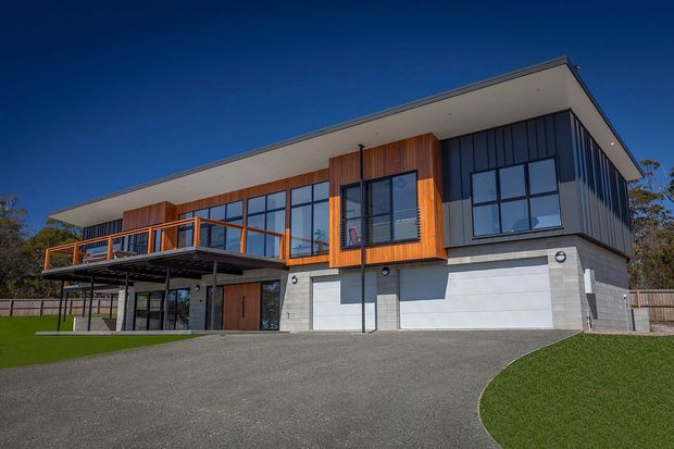 A contemporary two-storey residential home viewed from the front, featuring a broad low-pitched roof, dark vertical cladding, warm timber wall panels, extensive black-framed glazing, a projecting upper-level balcony, and a concrete lower level. The house is set on a landscaped suburban site with a curved driveway and lawn.